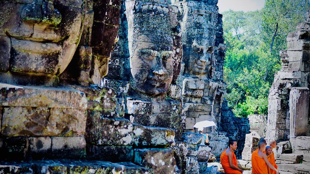 Ancient smiling stone faces of Bayon Temple overlooking Buddhist monks in orange robes at Angkor Thom, Siem Reap, Cambodia